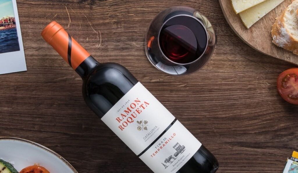 Vinho Tines de Roqueta Tempranillo. Vinho tinto Ramón Roqueta deitado sobre uma mesa de madeira. Ao lado uma taça transparente de vinho com vinho tinto. Uma tábua de queijos e pães ao lado, complementam a imagem.