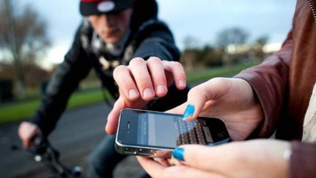 
Uma mulher está prestes a ter seu celular roubado por um ciclista que passa em alta velocidade, e estende a mão até o smartphone da vítima destruída. É dia, o ladrão tem o rosto borrado. O rosto da mulher não aparece.